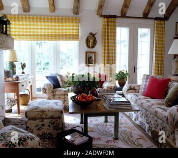 Yellow+white checked blind and curtains on French windows in a sunny country living room with floral sofas and ottoman Stock Photo