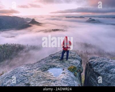 Hike with backpack and trekking sticks at the edge.  Woman visit dangerous and populat viewpoint above deep valley with thick mist. Stock Photo