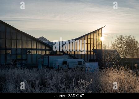 Magdeburg, Germany. 05th Dec, 2019. Behind the hypar dish, the sun rises. Construction vehicles and containers are standing in front of the multi-purpose hall. The listed building has been renovated since this week. Planned total costs: 17 million euros. According to the plan, the work will be completed in the summer of 2022. Credit: Stephan Schulz/dpa-Zentralbild/ZB/dpa/Alamy Live News Stock Photo