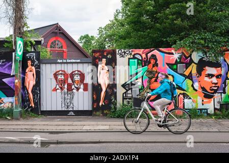 Christiania Denmark, view of a woman cycling past a colorful illustrated fence in the alternative Freetown area of Christiania, Copenhagen, Denmark Stock Photo