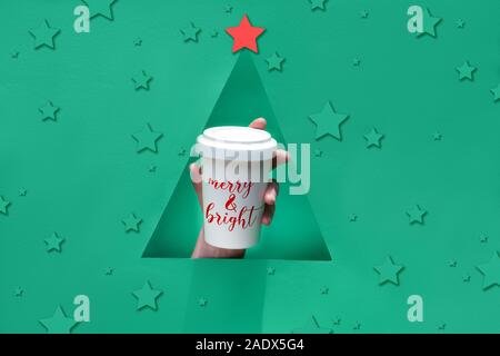 Stylish reusable eco coffee travel mug, bamboo cup with lid in hand through triangular paper hole in shape of Christmas tree. Green paper background, Stock Photo