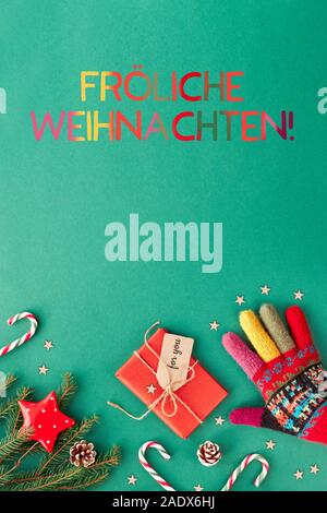 Text 'Frohliche Weihnachten' in German meaning 'Happy Christmas'. Xmas flat lay background in green and red on paper . Hand in wool glove, candy canes Stock Photo