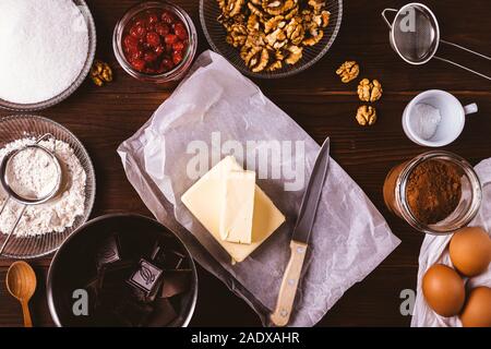 Sliced ​​butter among ingredients for baking homemade brownie, flour, eggs, cocoa, chocolate and delicious cherries and nuts additives on wooden table Stock Photo