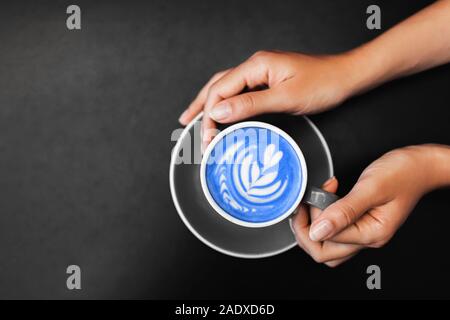 Cup of blue fresh matcha latte in woman hands on dark black metal table background. Empty place for text. Coffee addiction. View from above Stock Photo