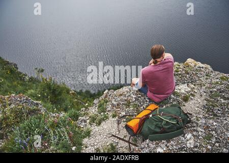 Traveller with backpack sitting on top of mountain enjoying view above the water surface. Traveling along mountains and coast, freedom and active Stock Photo