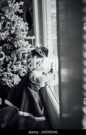 Baby girl looking out window next to Christmas tree Stock Photo