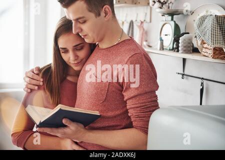 Hugs each other. Cute couple reading book together at home in the kitchen at daytime Stock Photo