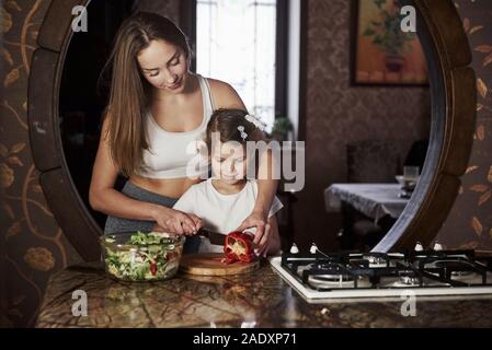 Tries to slice vegetable. Pretty young woman standing in the modern kitchen near gas stove and teach daughter how to prepare food