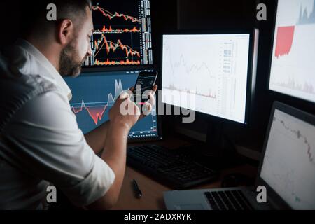 New modern digital technologies. Bearded man in white shirt works by using smartphone in the office with multiple computer screens in index charts Stock Photo
