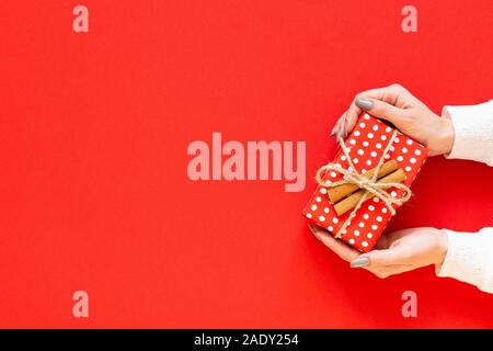 Girl holds red gift box in polka dots with cinnamon on a red background, merry christmas and happy new year concept, flat lay, top view Stock Photo