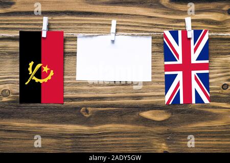 Hanging flags of Angola and United Kingdom attached to rope with clothes pins with copy space on white note paper on wooden background.Diplomatic rela Stock Photo