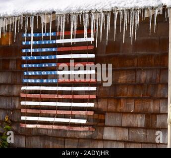 American flag painted on a snow fence hangs from a shed eave trimmed with icicles. Stock Photo