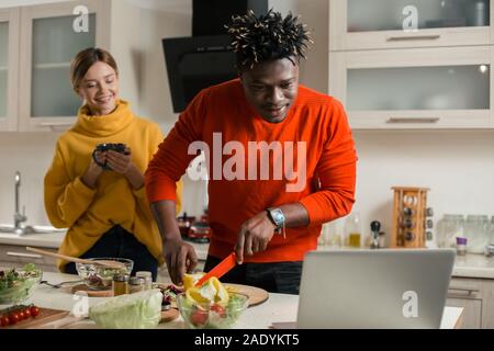 Smiling man cutting paprika and looking at the screen of laptop Stock Photo