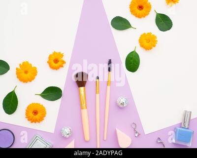 Fresh flower and green leaves on white papers and natural cosmetics, and makeup tools and brushes, mirror isolated on a purple background. Frame made Stock Photo
