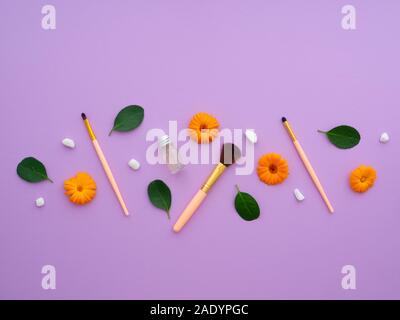 Makeup brushes, everyday make-up tools. Closeup cosmetic essentials Shampoo, green leaves, orange flowers, silver balls, on a purple background. Stock Photo