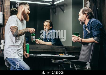 Two men smiling while drinking beer in barbershop Stock Photo