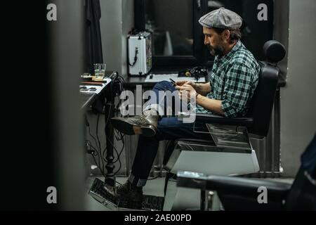 Full length of bearded man sitting in barber chair and looking at smartphone Stock Photo