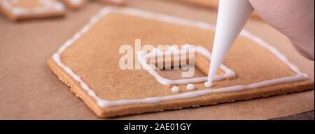 Woman is decorating gingerbread cookies house with white frosting icing cream topping on wooden table background, baking paper in kitchen, close up, m Stock Photo