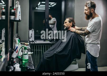 Middle aged man staying calm and barber putting hairdressing gown on him Stock Photo
