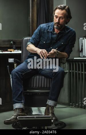 Calm man in jeans clothes sitting in barbershop chair and thinking Stock Photo