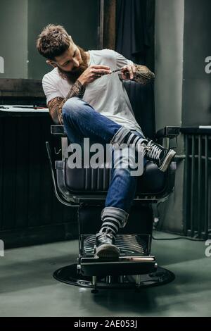 Bearded man sitting in barber armchair and touching razors Stock Photo