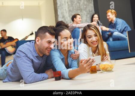 Group of friends having party indoors fun together lying on the floor taking selfies Stock Photo