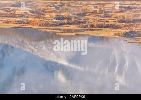 Colorful autumn view with golden larch trees on the shore of the lake, hoarfrost on the grass, fog and reflection of snowy mountains in the water Stock Photo