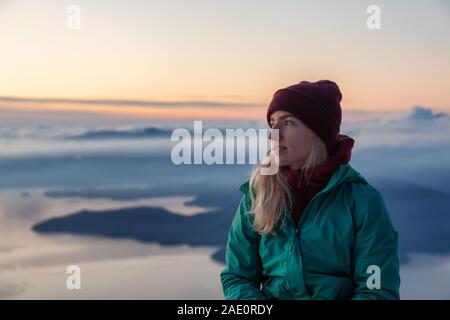 Adventurous Blond Caucasian Girl sitting on top of a snow covered mountain during a colorful winter sunset. Taken on St Mark's Summit, North of Vancou Stock Photo