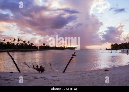 North Male Atoll, Maldives - November 23, 2019: A happy adult couple relaxes in a hammock over the water at a resort in the Maldives during sunset Stock Photo