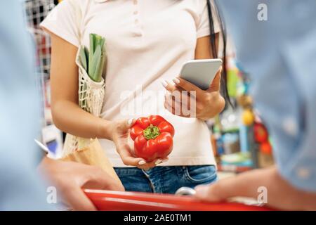 Daily Shopping. Couple in the supermarket with cart together holding bell pepper browsing smartphone close-up Stock Photo