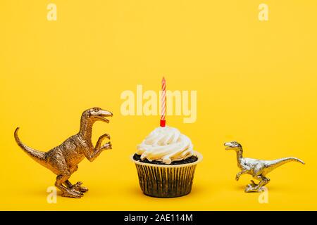 Toy dinosaurs beside cupcake with candle on yellow background Stock Photo