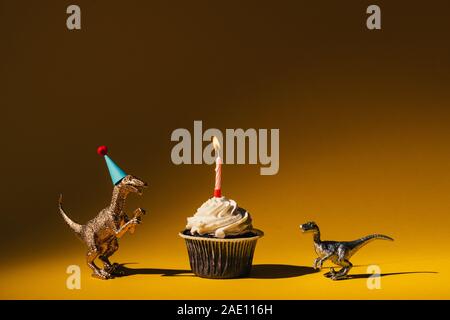 Toy dinosaurs with party cap and cupcake with burning candle on orange background Stock Photo