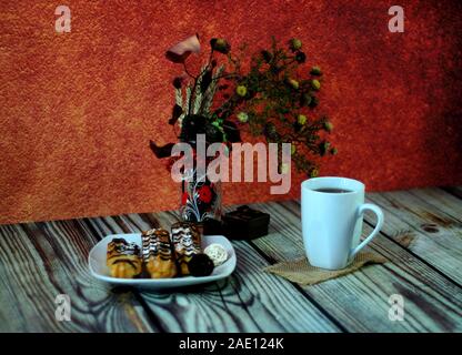 Warm still life, a plate with fresh eclairs, a white ceramic mug with tea on a napkin and a glass vase with an autumn herbarium. Close-up. Stock Photo