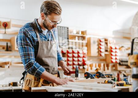 selective focus of carpenter in goggles holding hammer near wooden dowel in workshop Stock Photo