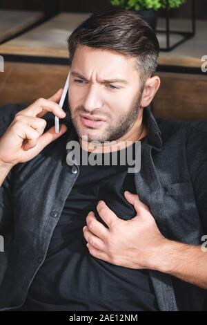 handsome man having heart attack and talking on smartphone in apartment Stock Photo