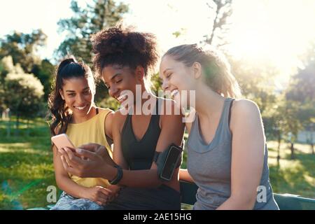 Smiling female diverse friends looking at mobile phone smiling and having fun in the morning at the park - friends laughing together Stock Photo