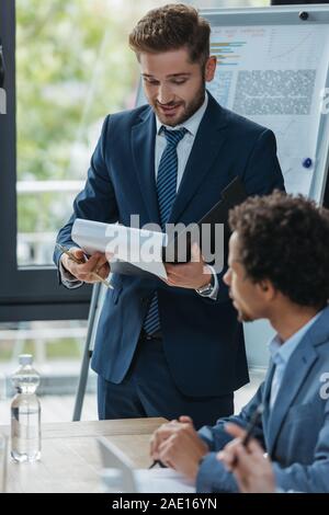 selective focus of young businessman reading document at business meeting with multicultural colleagues Stock Photo