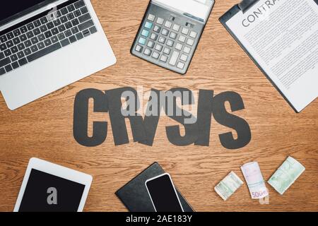 top view of word crisis near digital devices, clipboard with contract, cash rolls of dollar, euro and ruble banknotes on wooden desk Stock Photo