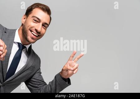 handsome and smiling businessman in suit showing peace sign isolated on grey Stock Photo