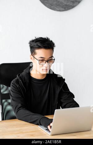 asian hacker in glasses sitting at table and using laptop Stock Photo