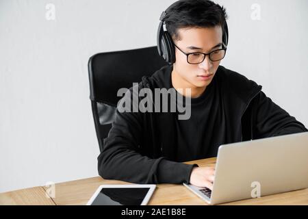 asian hacker in headphones using laptop and sitting at table Stock Photo