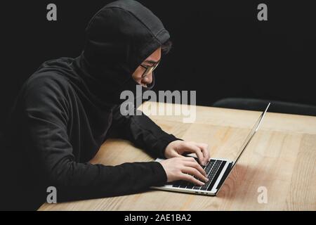 high angle view of asian hacker with obscured face using laptop and sitting at table Stock Photo