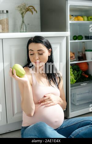 happy pregnant woman holding apple while sitting on floor in kitchen near opened fridge Stock Photo