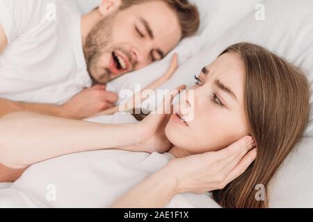 selective focus of woman plugging ears with hands while lying in bed near snoring husband Stock Photo