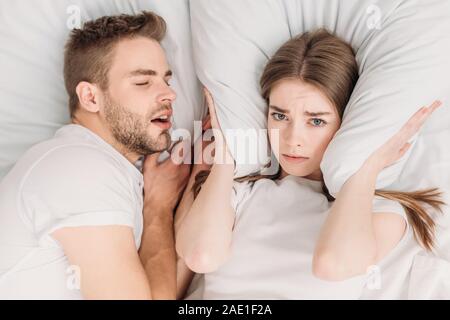 top view of woman plugging ears with pillow while lying in bed near snoring husband Stock Photo