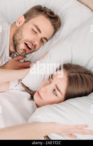 awakened woman plugging ears with pillow while lying in bed near snoring husband Stock Photo