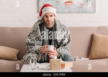 sick man in santa hat looking at camera while holding cup of warm drink Stock Photo