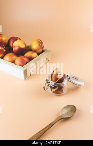 Glass jar full of slices of nectarines, a wooden spoon and a box full of peaches in the back , on a peachy colored background Stock Photo
