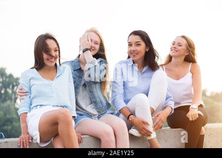 Four attractive women in summer clothes sitting at concrete border near river after studing in college. Young student girls having fun Stock Photo