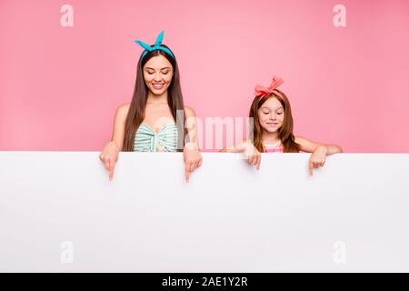 Portrait of curious woman with her schoolkid having brunette red long hairdo show with index finger ads wear bright headbands isolated over pink Stock Photo